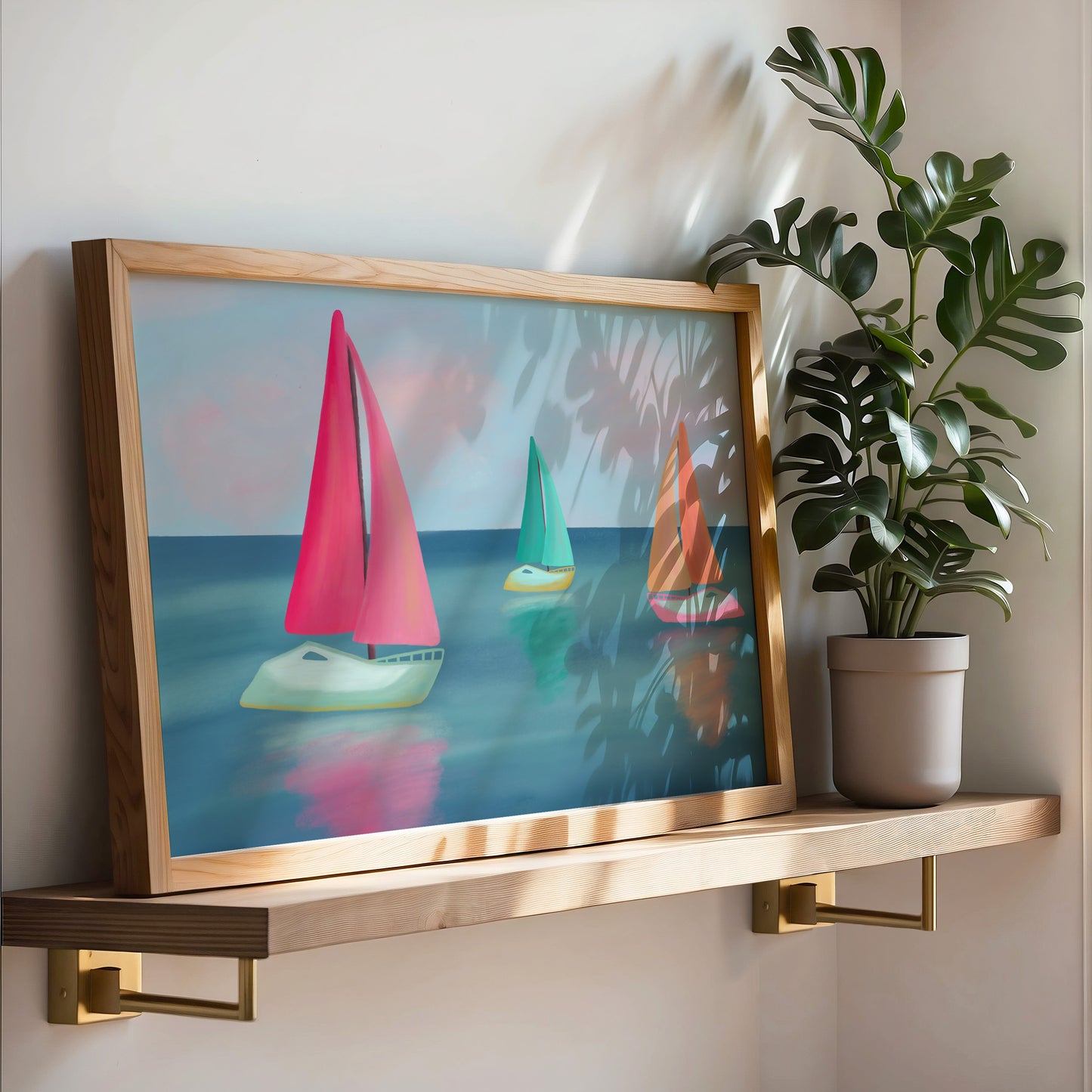 A framed art print featuring three colorful sailboats on a blue sea, styled on a wooden floating shelf next to a potted monstera plant.