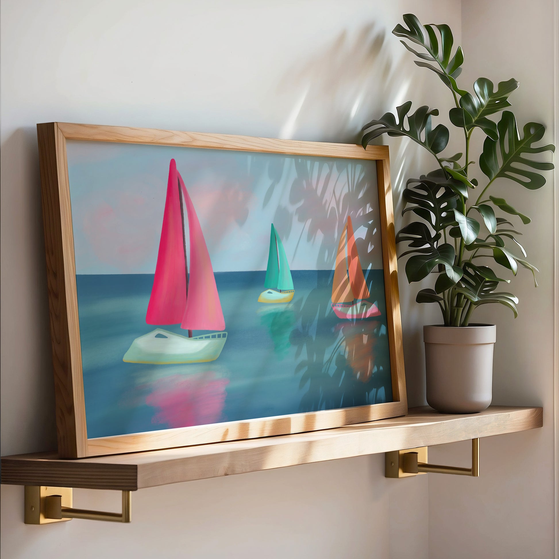A framed art print featuring three colorful sailboats on a blue sea, styled on a wooden floating shelf next to a potted monstera plant.